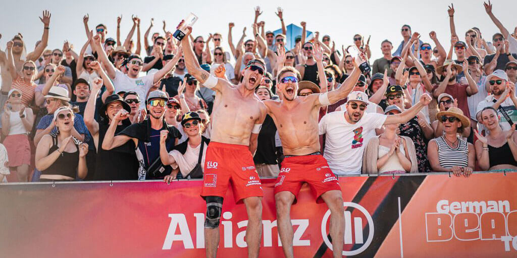 Beachvolleyballer Bennet Poniewaz und sein Teamkollege ohne Oberkörper in roten Shorts jubeln mit erhobenen Armen und Trophäen vor einer großen, klatschenden Zuschauermenge bei der German Beach Tour.