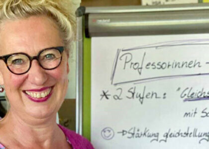 Bettina Belker steht lächelnd neben einem Whiteboard, auf dem Notizen zum "Professorinnen-Programm" und "Gleichstellung" stehen. Sie trägt eine Brille und eine pinkfarbene Jacke.