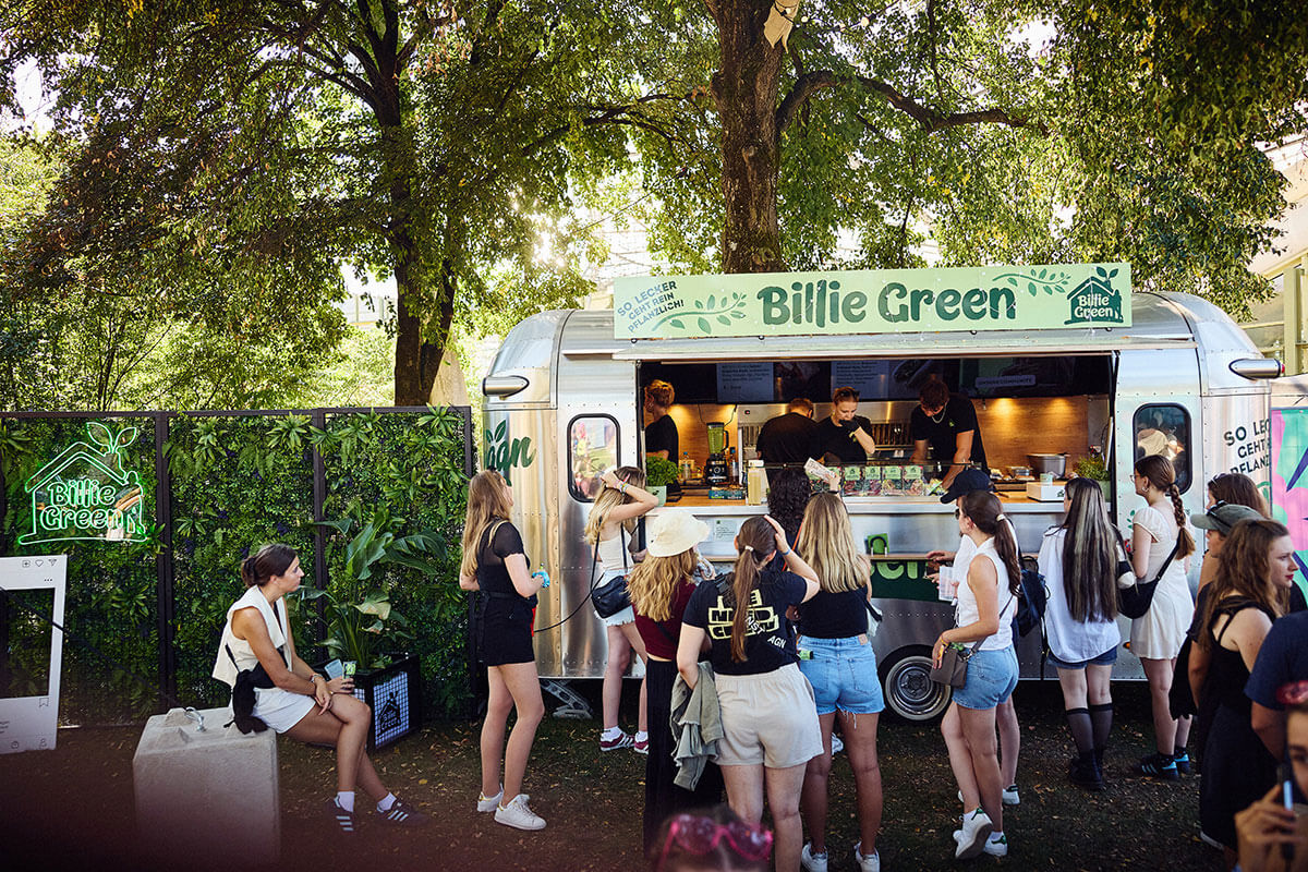 Warteschlange von Menschen vor einem silbernen Billie-Green-Foodtruck im Retro-Design unter Bäumen.