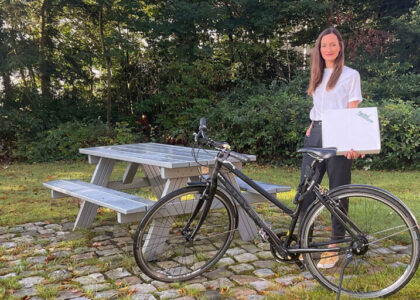 Maren Weitner steht lächelnd neben einem Fahrrad vor einer Picknickbank im Grünen und hält ein Paket im Arm.