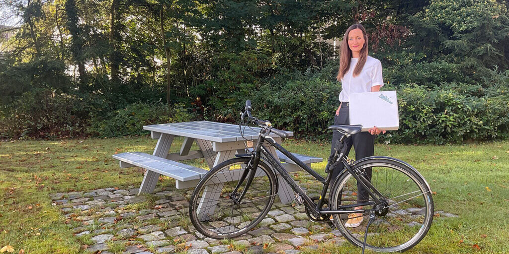 Maren Weitner steht lächelnd neben einem Fahrrad vor einer Picknickbank im Grünen und hält ein Paket im Arm.