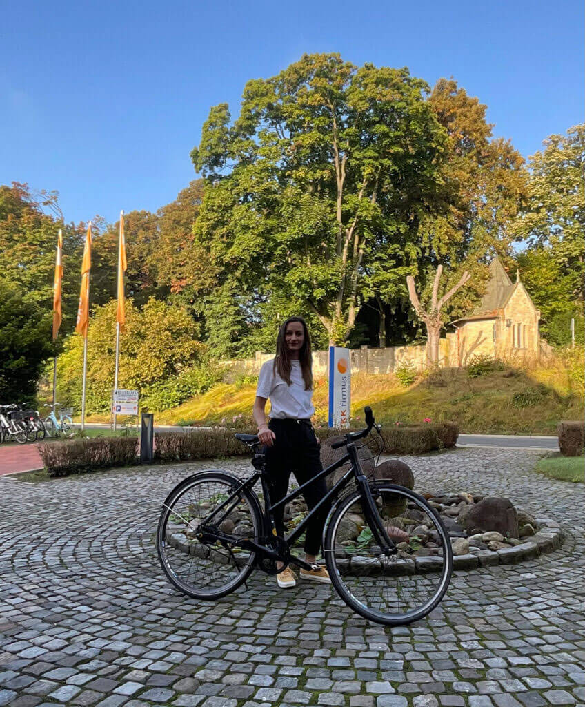 Maren Weitner steht lächelnd mit einem schwarzen Fahrrad auf einem gepflasterten Platz vor einem Gebäude der BKK firmus.