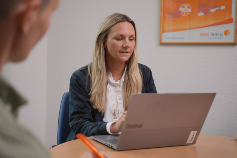 Blonde Frau sitzt an einem Tisch vor geöffnetem Laptop und spricht mit einer Person im Vordergrund.