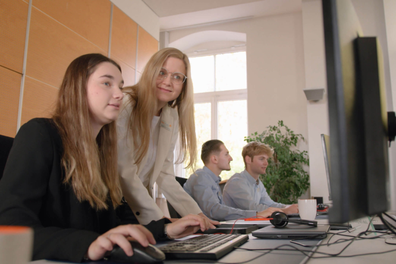 Blonde Frau im hellen Blazer beugt sich lächelnd zu einer Kollegin am PC-Arbeitsplatz.