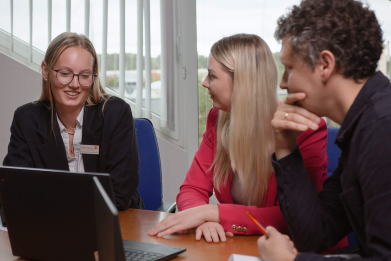 Drei Personen sitzen an einem Tisch um einen Laptop, eine Frau mit Namensschild 