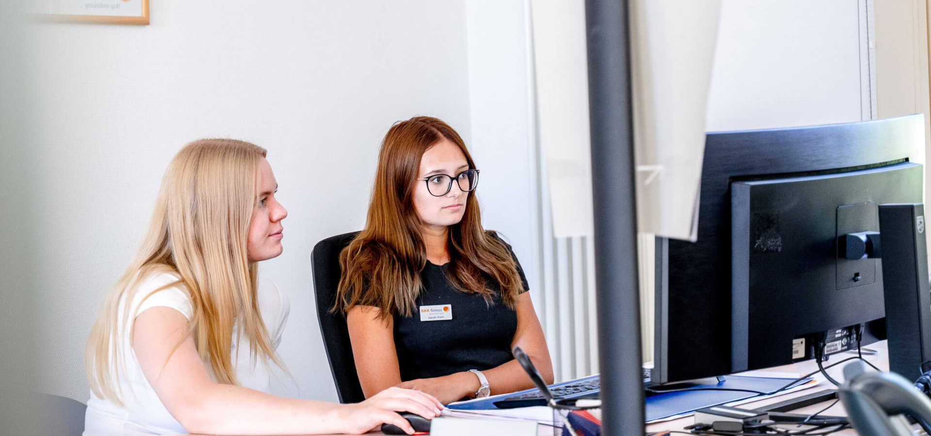 Zwei Frauen sitzen nebeneinander an einem Schreibtisch und schauen gemeinsam auf einen großen Monitor.