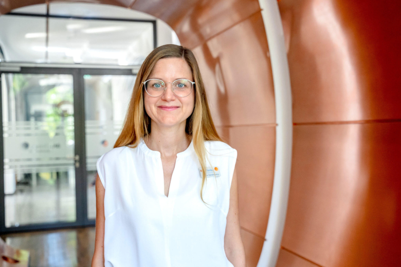 Blonde Frau mit Brille und weißem Oberteil lächelt vor einer geschwungenen, kupferfarbenen Wand.