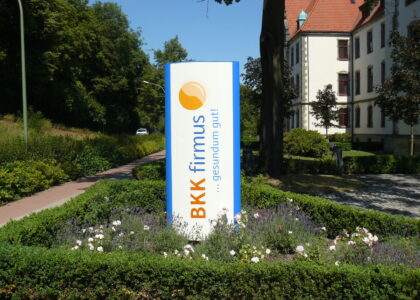 Aufgestellte Infosäule der Krankenkasse BKK firmus in einem gepflegten Grünbeet vor einem historischen Gebäude.