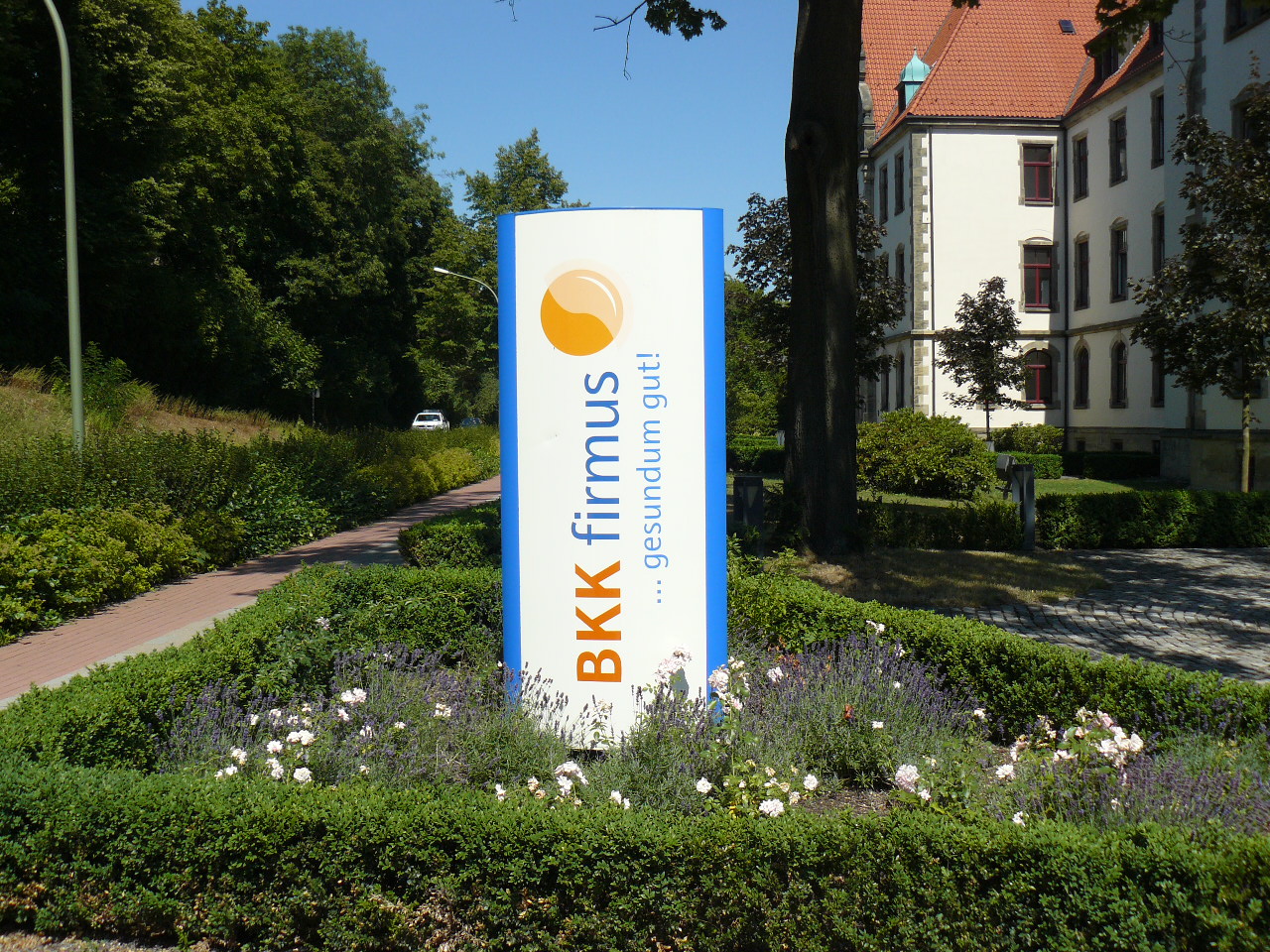 Aufgestellte Infosäule der Krankenkasse BKK firmus in einem gepflegten Grünbeet vor einem historischen Gebäude.