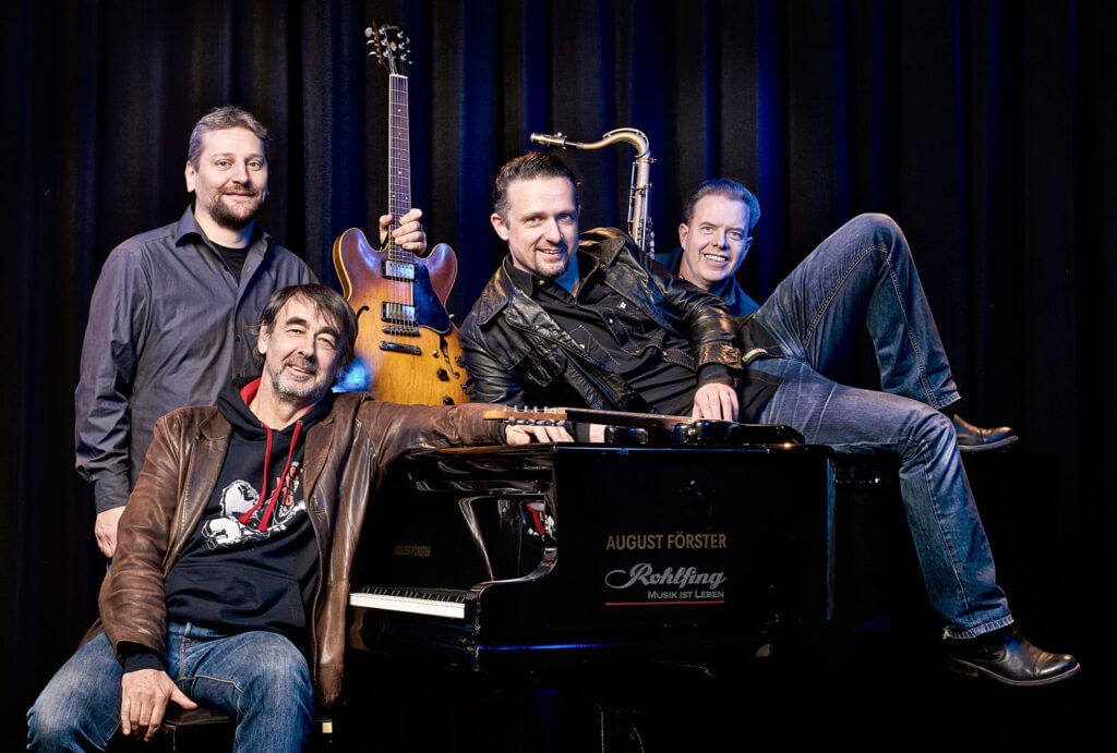 Vierköpfige Männerband posiert lächelnd mit Gitarre und Saxophon an und auf einem schwarzen Flügel vor dunklem Vorhang.