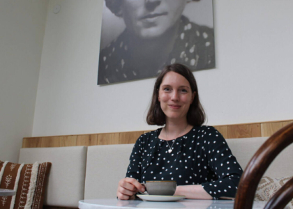 Eine Frau im gepunkteten Oberteil sitzt an einem Cafétisch und hält eine Kaffeetasse; im Hintergrund hängt ein großes Schwarz-Weiß-Porträt.