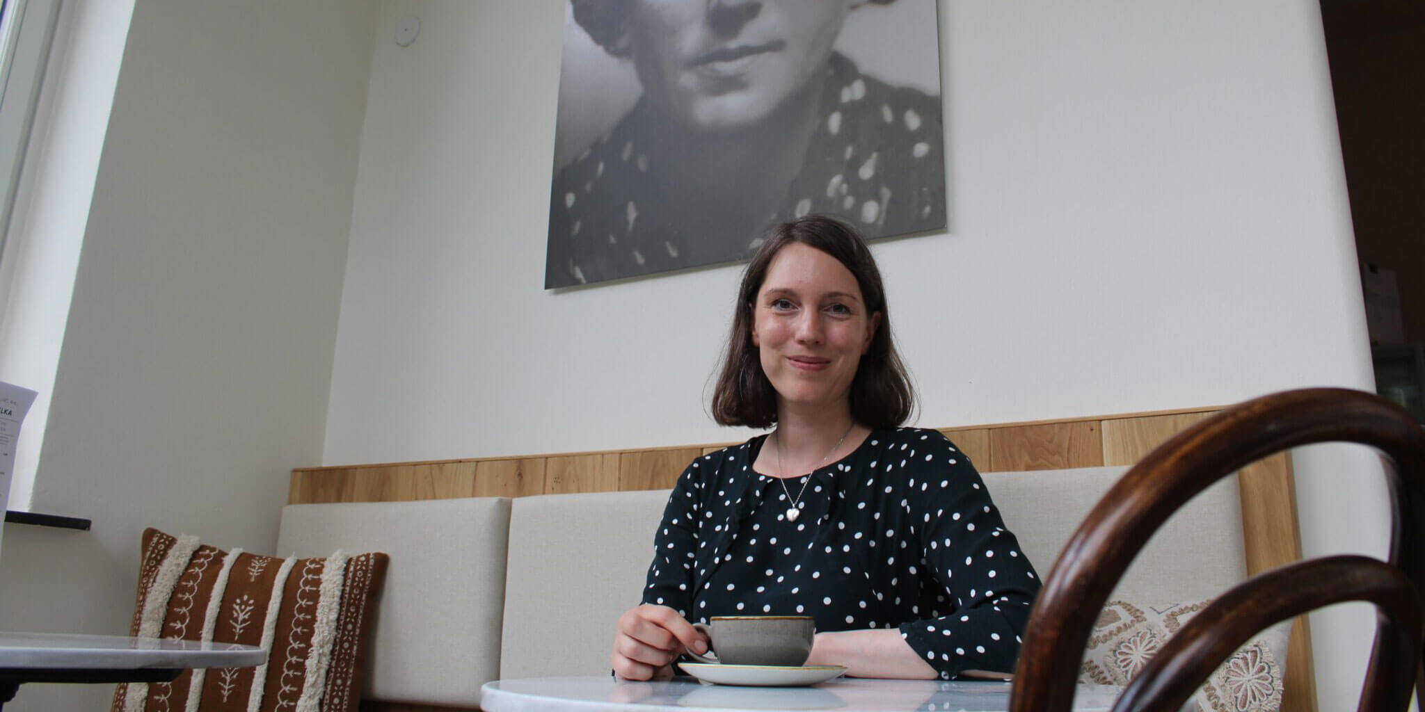Eine Frau im gepunkteten Oberteil sitzt an einem Cafétisch und hält eine Kaffeetasse; im Hintergrund hängt ein großes Schwarz-Weiß-Porträt.