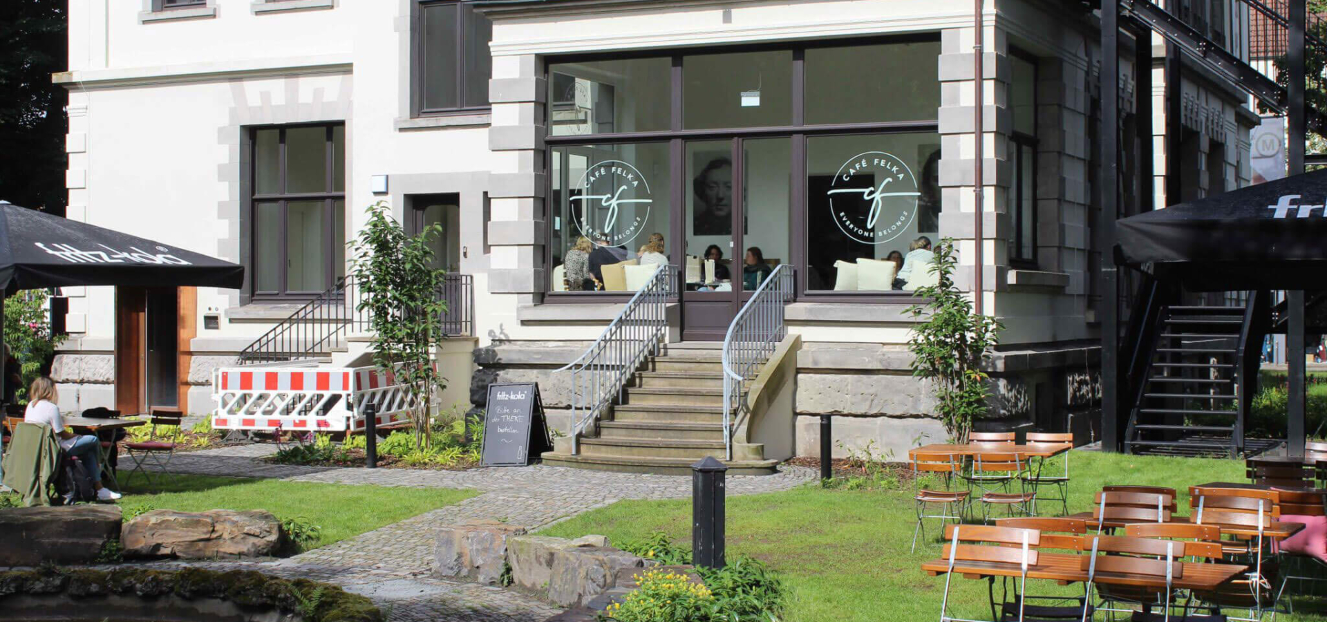 Außenansicht des Café Felka in einer Altbauvilla mit Terrasse, grünem Rasen und hölzernen Sitzgruppen im Vordergrund.