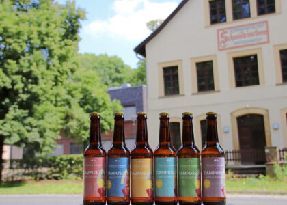 Sechs Bierflaschen mit bunten Etiketten der Campusbier-Jubiläumsedition stehen aufgereiht vor einem historischen Gebäude.