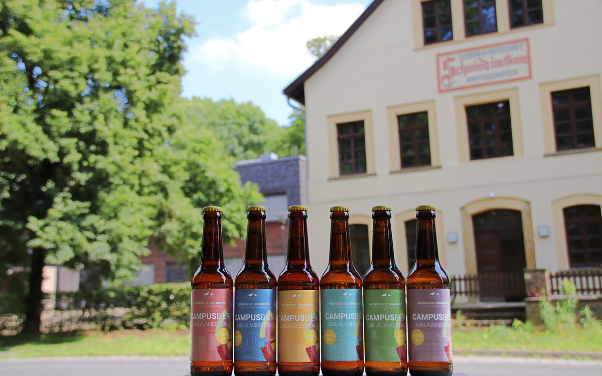 Sechs Bierflaschen mit bunten Etiketten der Campusbier-Jubiläumsedition stehen aufgereiht vor einem historischen Gebäude.