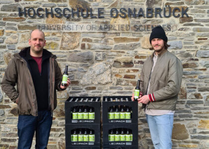 Zwei Männer stehen mit Bierflaschen und Bierkästen vor einer Steinmauer mit dem Schriftzug Hochschule Osnabrück.