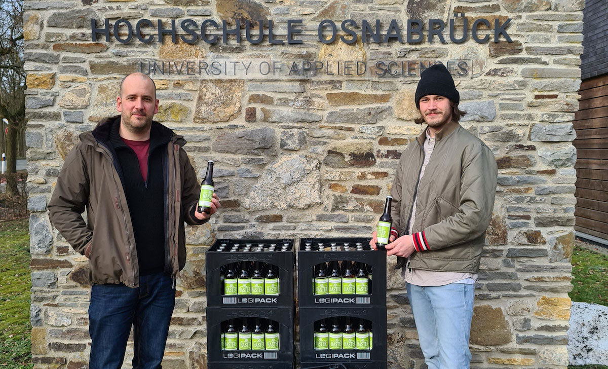 Zwei Männer stehen mit Bierflaschen und Bierkästen vor einer Steinmauer mit dem Schriftzug Hochschule Osnabrück.