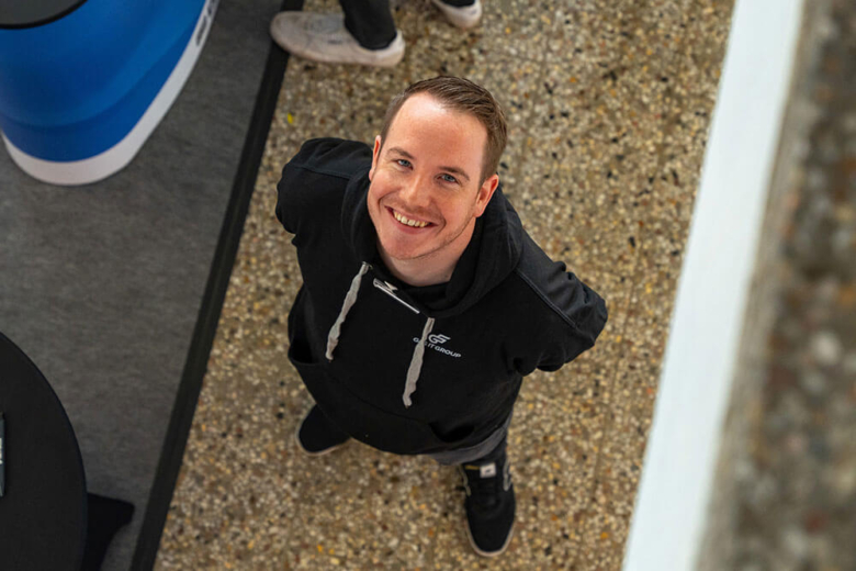 Ein junger Mann im schwarzen Hoodie blickt lächelnd von unten nach oben in die Kamera.