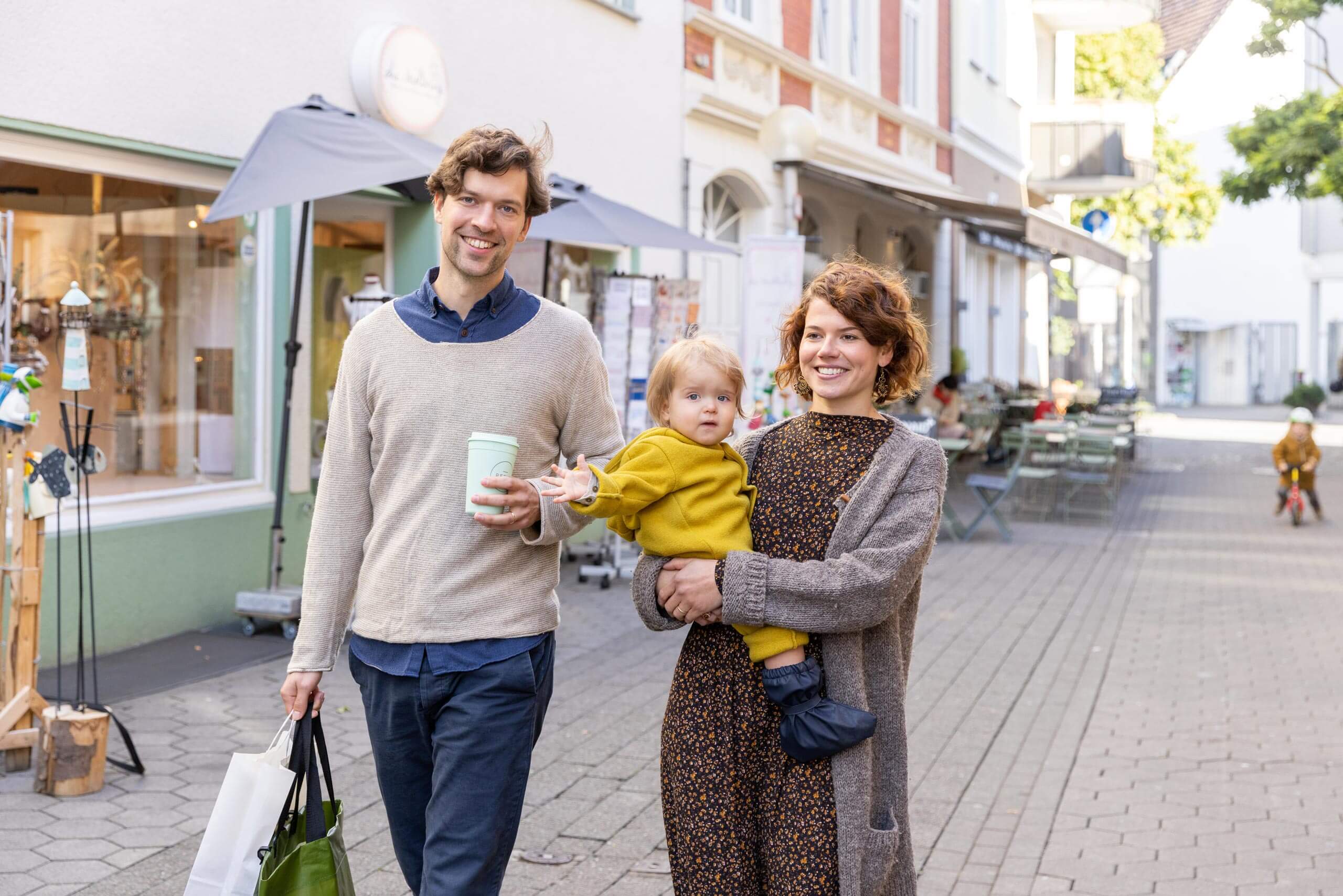 Ein Paar geht lächelnd durch eine Fußgängerzone, während die Frau ein winkendes Kleinkind auf dem Arm trägt und der Mann Taschen und Kaffee hält.