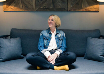 Corinna Baethge sitzt lächelnd im Schneidersitz auf einem gemütlichen, dunklen Sofa.
