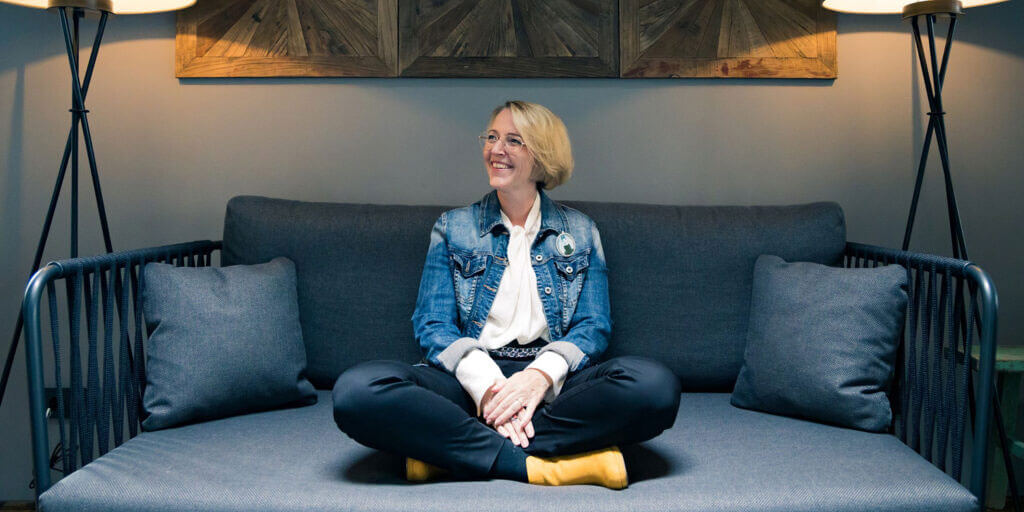 Corinna Baethge sitzt lächelnd im Schneidersitz auf einem gemütlichen, dunklen Sofa.