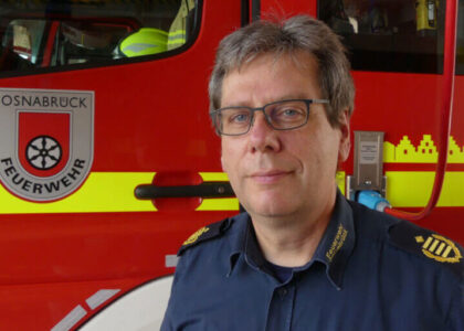 Porträt von Dietrich Bettenbrock mit Brille in einer dunkelblauen Uniform der Feuerwehr Osnabrück, der vor der roten Seitenwand eines Feuerwehrfahrzeugs steht.