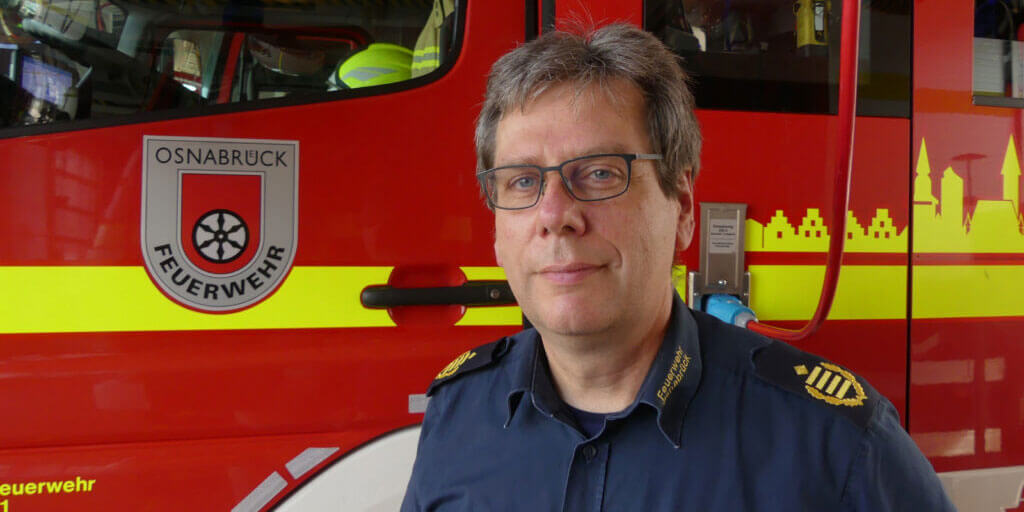 Porträt von Dietrich Bettenbrock mit Brille in einer dunkelblauen Uniform der Feuerwehr Osnabrück, der vor der roten Seitenwand eines Feuerwehrfahrzeugs steht.