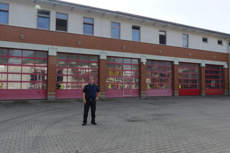 Dietrich Bettenbrock in dunkelblauer Feuerwehruniform steht auf dem gepflasterten Vorplatz einer Feuerwache vor einer Reihe von geschlossenen, roten Garagentoren mit Glasscheiben.