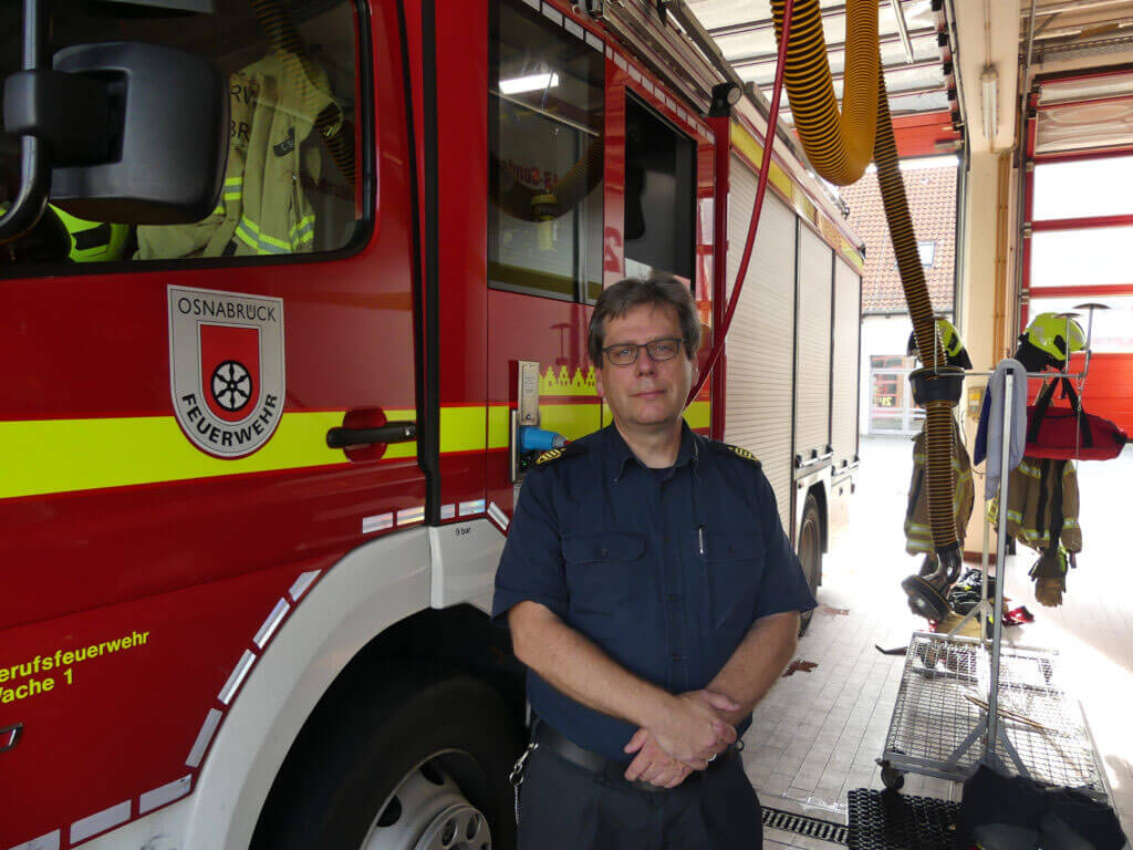 Feuerwehrmann Dietrich Bettenbrock in dunkelblauer Uniform steht mit verschränkten Händen in einer Fahrzeughalle neben der roten Fahrertür eines Einsatzwagens der Berufsfeuerwehr Osnabrück.