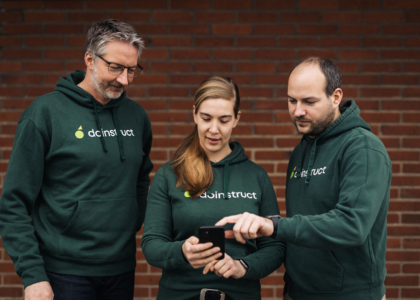Drei Personen in grünen doinstruct-Hoodies stehen vor einer Backsteinwand und schauen gemeinsam auf ein Smartphone.
