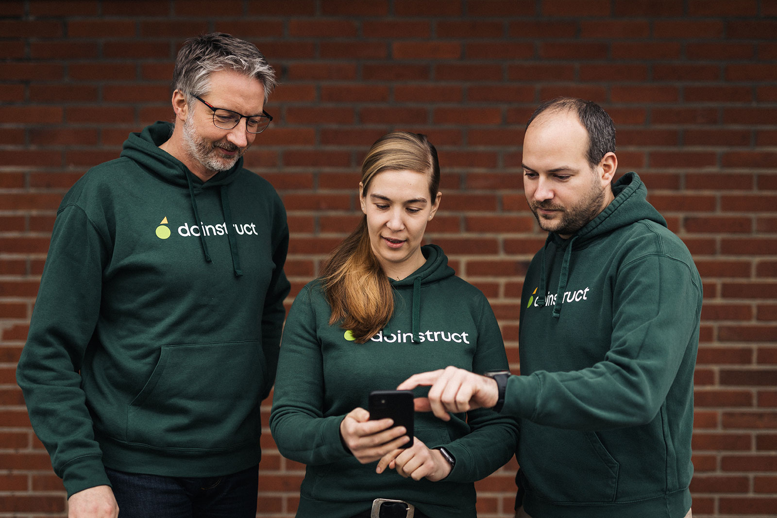 Drei Personen in grünen doinstruct-Hoodies stehen vor einer Backsteinwand und schauen gemeinsam auf ein Smartphone.