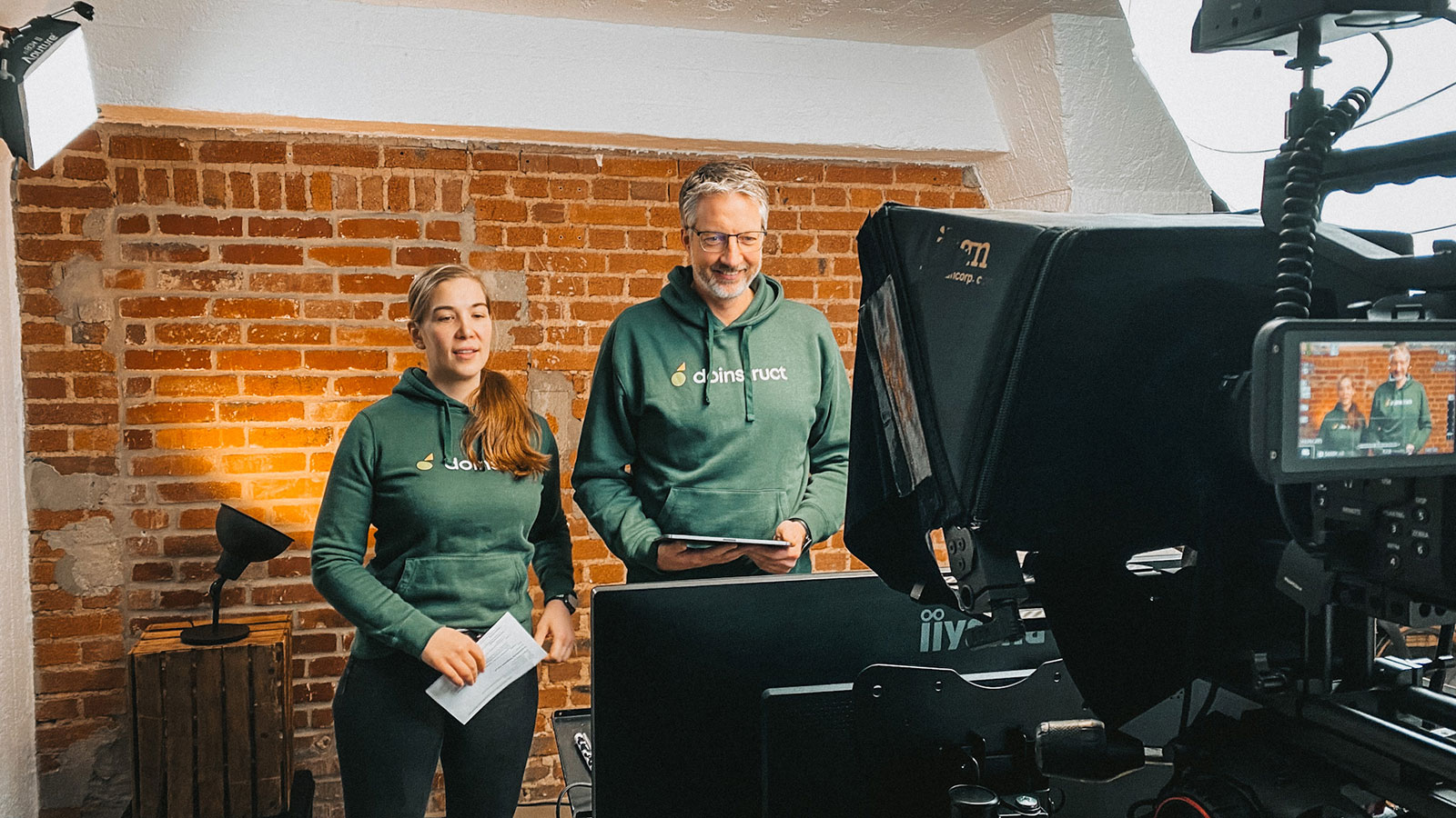 Eine Frau und ein Mann in grünen Hoodies stehen vor einer Backsteinwand hinter einer Studiokamera.