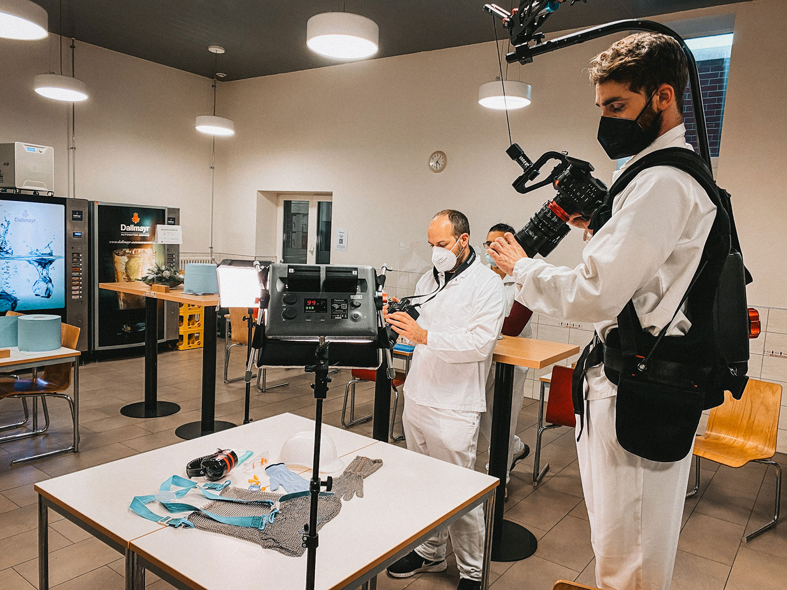 Kamerateam in weißer Schutzkleidung und FFP2-Masken filmt Schutzausrüstung auf einem Tisch in einem Pausenraum.