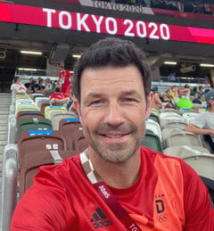 Selfie von Dr. Casper Grim im roten T-Shirt des Team Deutschland auf den Zuschauerrängen eines Stadions mit der Anzeige „TOKYO 2020“ im Hintergrund.