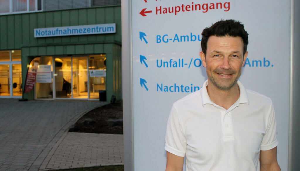 Porträt von Dr. Casper Grim im weißen Poloshirt vor einem Hinweisschild des Notaufnahmezentrums einer Klinik.
