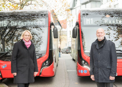 Eine Frau und ein Mann stehen lächelnd vor zwei rot-schwarzen Elektro-Stadtbussen der Verkehrsbetriebe an einer Haltestelle.