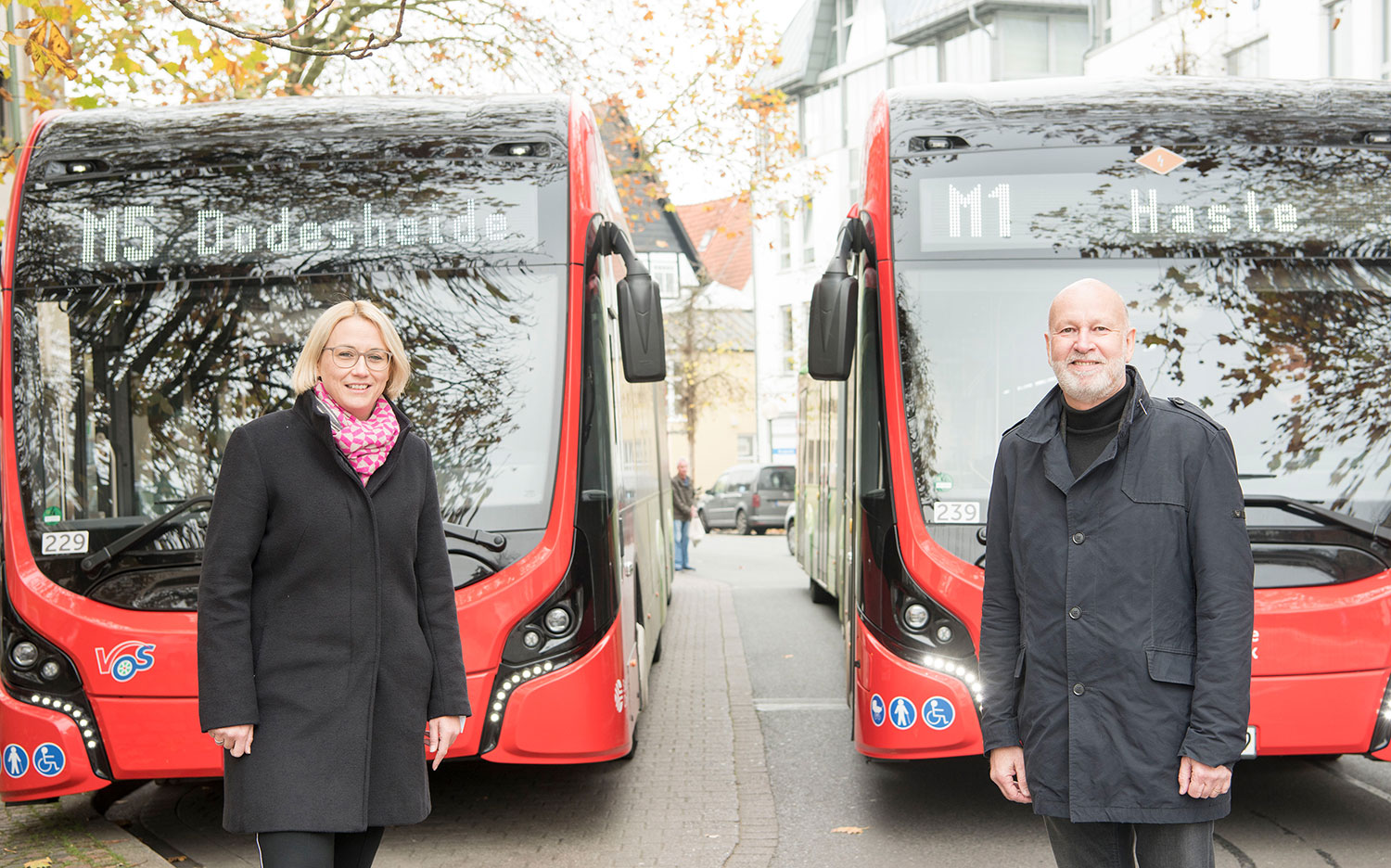 Eine Frau und ein Mann stehen lächelnd vor zwei rot-schwarzen Elektro-Stadtbussen der Verkehrsbetriebe an einer Haltestelle.