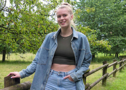 Porträtfoto von Emily Enochs im Freien. Sie trägt ein grünes Oberteil und eine offene Jeansjacke und lehnt an einem Holzzaun. Im Hintergrund sind Bäume und ein Feld zu sehen.