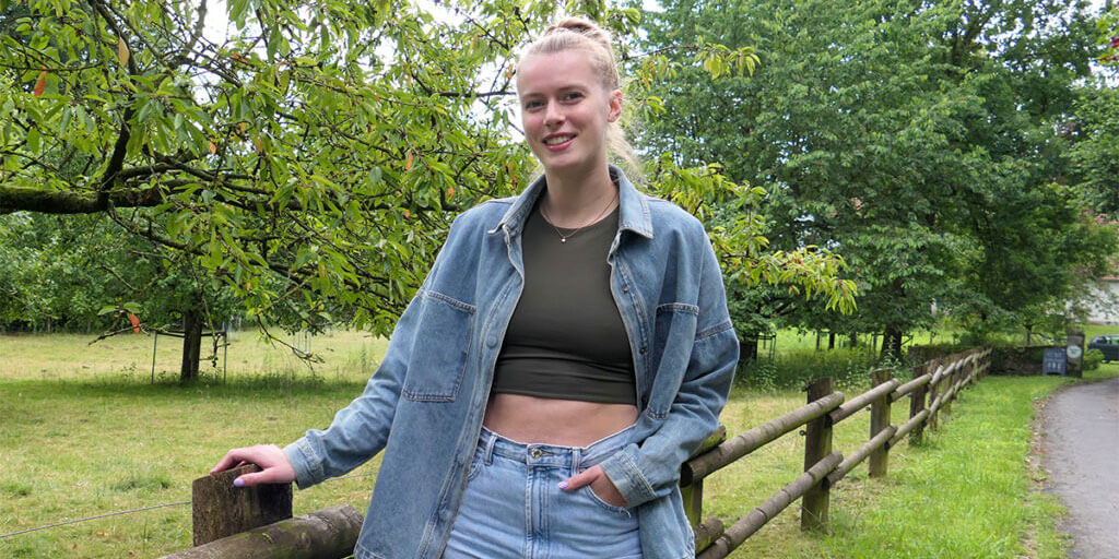 Porträtfoto von Emily Enochs im Freien. Sie trägt ein grünes Oberteil und eine offene Jeansjacke und lehnt an einem Holzzaun. Im Hintergrund sind Bäume und ein Feld zu sehen.