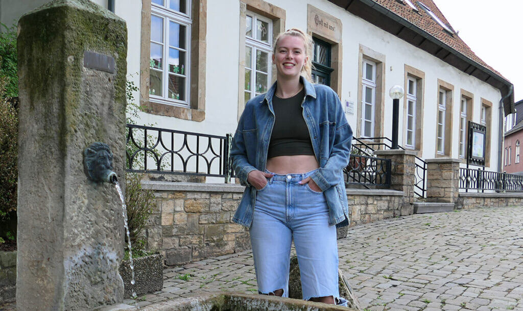 Ganzkörperfoto von Emily Enochs im Freien. Sie steht lächelnd in einer gepflasterten Straße vor einem alten, weißen Haus mit Steinsockel. Links von ihr steht ein steinerner Brunnen mit einem Löwenkopf.