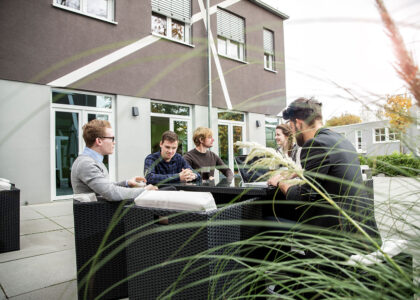 Eine Gruppe von jungen Erwachsenen sitzt zusammen an einem Tisch auf einer Terrasse vor einem modernen Bürogebäude.