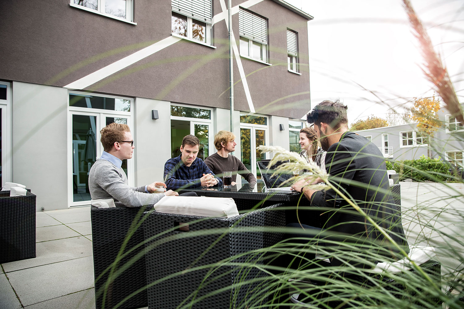 Eine Gruppe von jungen Erwachsenen sitzt zusammen an einem Tisch auf einer Terrasse vor einem modernen Bürogebäude.