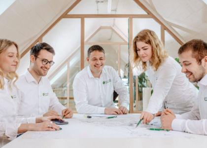 Fünf Personen in weißen Hemden/Blusen mit Firmenlogo besprechen Pläne an einem großen, hellen Tisch im Dachgeschoss.