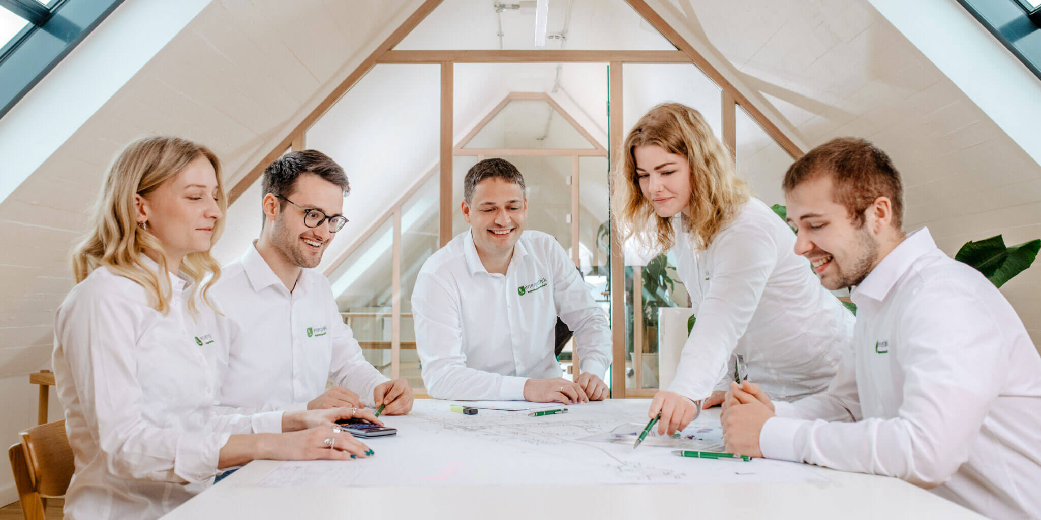 Fünf Personen in weißen Hemden/Blusen mit Firmenlogo besprechen Pläne an einem großen, hellen Tisch im Dachgeschoss.