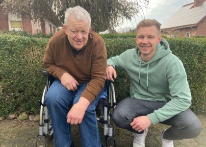 Ein älterer Mann im Rollstuhl und ein jüngerer Mann in Hocke lächeln gemeinsam vor einer Hecke.