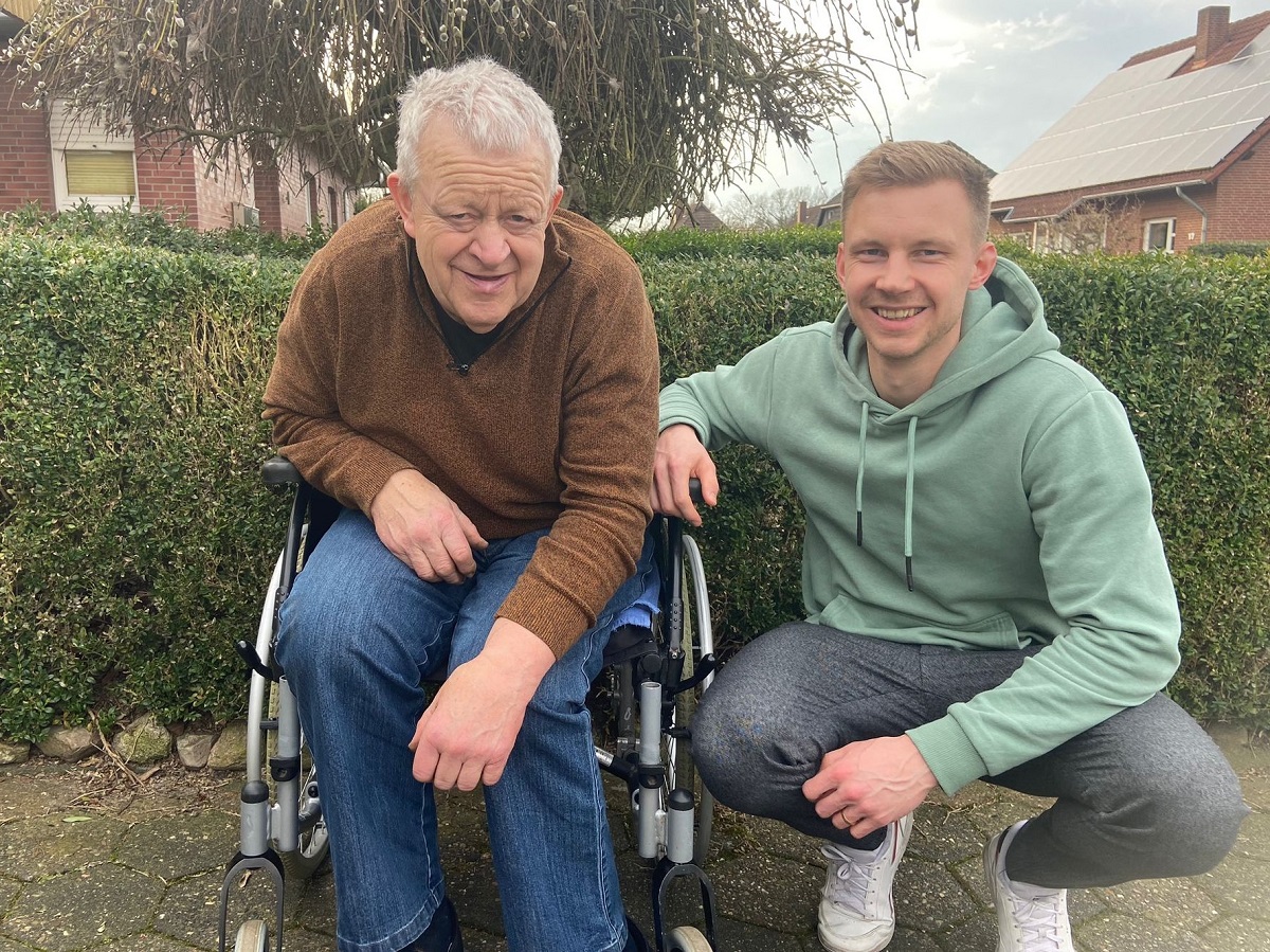 Ein älterer Mann im Rollstuhl und ein jüngerer Mann in Hocke lächeln gemeinsam vor einer Hecke.