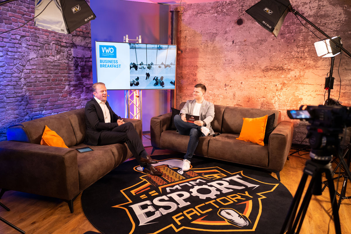 Zwei Männer im Anzug führen ein Gespräch auf braunen Sofas in einem Studio der eSport Factory mit Kamera und Bildschirm.