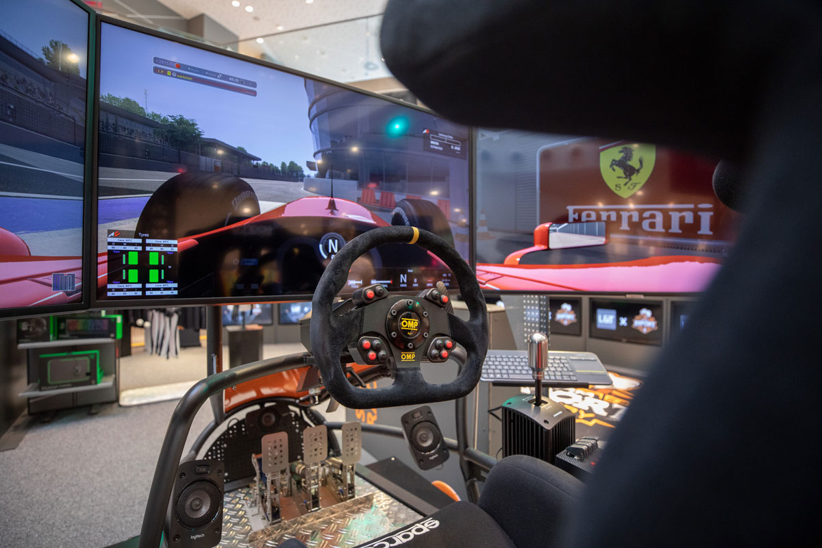Ein Rennsimulator-Cockpit mit drei Bildschirmen, die eine virtuelle Rennstrecke anzeigen, einem Lenkrad, Pedalen und einem Schalthebel.