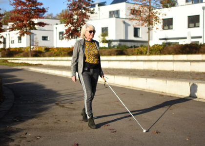 Eine ältere Frau mit Sonnenbrille und Blindenstock geht an einem sonnigen Tag auf einem gepflasterten Weg spazieren.