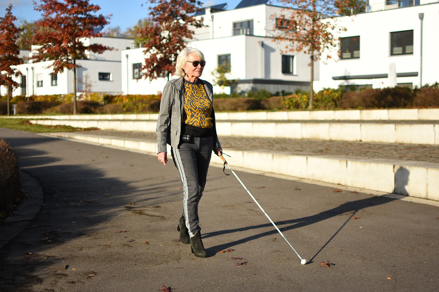Eine ältere Frau mit Sonnenbrille und Blindenstock geht an einem sonnigen Tag auf einem gepflasterten Weg spazieren.